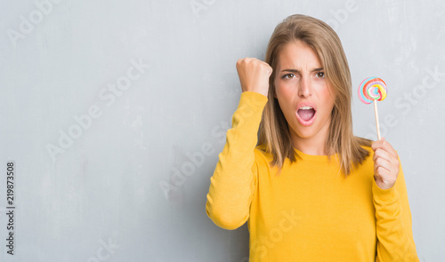 Beautiful young woman over grunge grey wall eating lollipop candy annoyed and frustrated shouting with anger, crazy and yelling with raised hand, anger concept