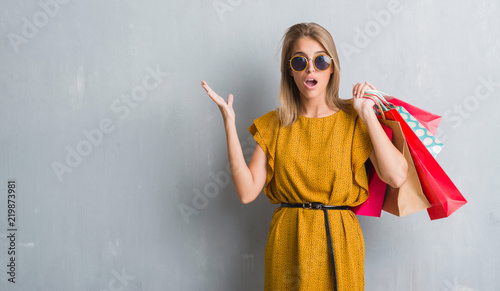 Beautiful young woman over grunge grey wall holding shopping bags on sales scared in shock with a surprise face, afraid and excited with fear expression