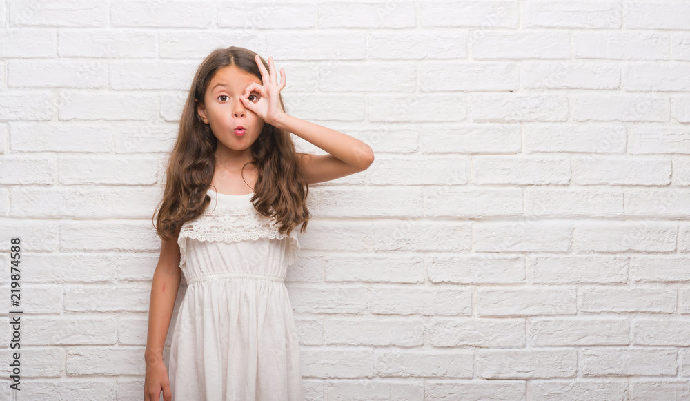 Young hispanic kid over white brick wall doing ok gesture shocked with surprised face, eye looking through fingers. Unbelieving expression. Young hispanic kid over white brick wall doing ok gesture shocked with surprised face, eye looking through fingers. Unbelieving expression.