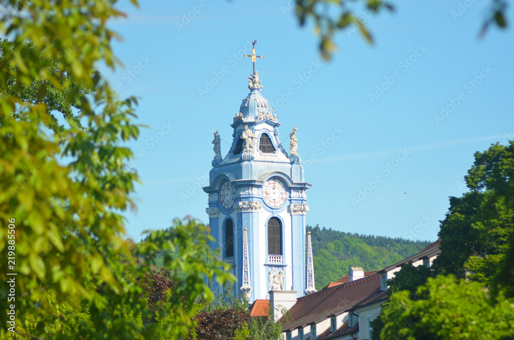 An ornate blue steeple rises against a blue sky. The roofs of the village. peek above the trees. A forest covered mountain is in the background.