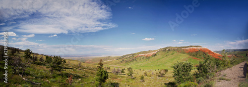 A panorama of hilly green pasture land dotted with trees and red rock along the Chief Joseph scenic highway in Montana