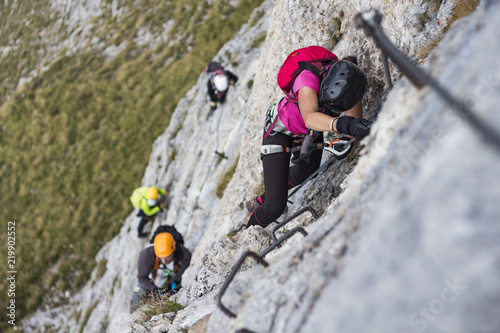 Woman and group of other people climbing on steep rock face on via ferrata. Climbers on via ferrata climbing route. Alpine ferrata ascent to summit. Summer adventure mountain activity. photo