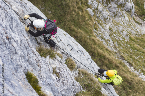 Woman and group of other people climbing on steep rock face on via ferrata. Climbers on via ferrata climbing route. Alpine ferrata ascent to summit. Summer adventure mountain activity.