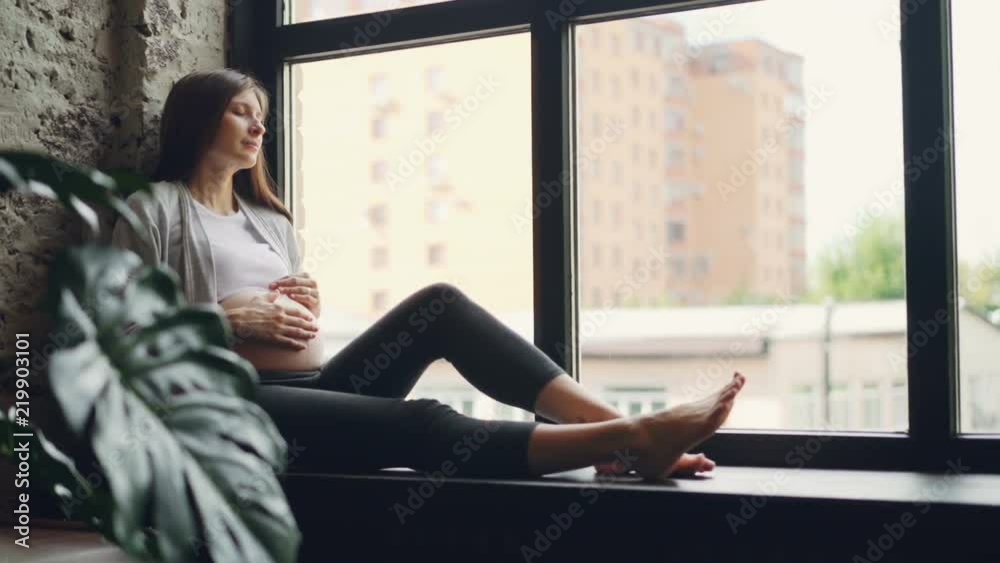 Attractive pregnant lady is sitting on window sill and caressing her baby bump, she is looking outside then closing her eyes and resting. Modern interiors and pregnancy concept. Attractive pregnant lady is sitting on window sill and caressing her baby bump, she is looking outside then closing her eyes and resting. Modern interiors and pregnancy concept.