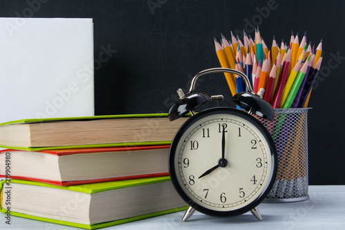 background of sets of office items and a black alarm clock on colorful books, next to a glass with pencils, there is a place for an inscription