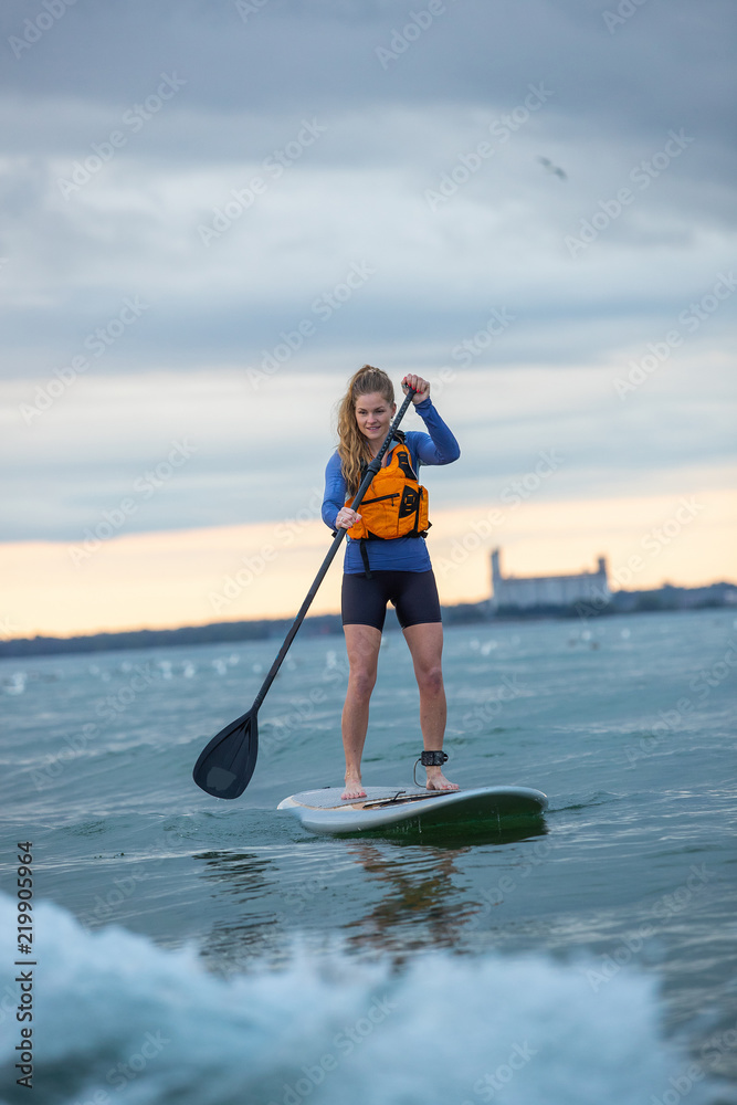 A beautiful woman paddle boards near a lighthouse in Ontario, Canada at sunrise or sunset