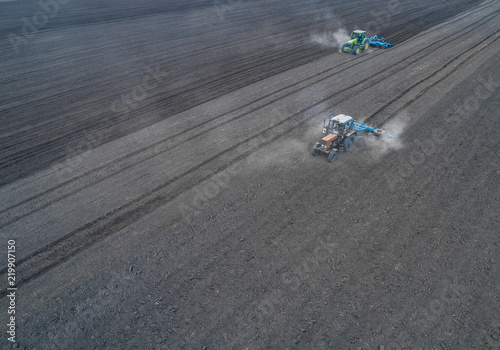 Aerial view of two blue tractors plows the earth in field on a summer day against a black earth background. Agriculture. Two tractors travel one after another along the black field