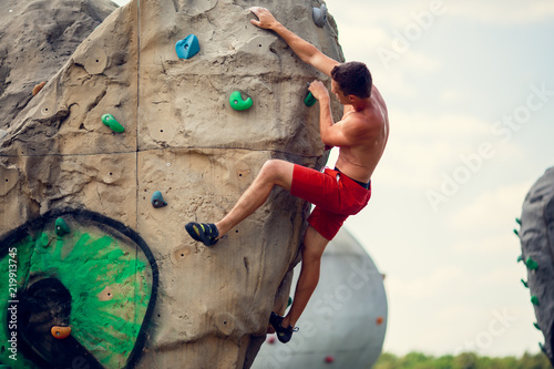 Photo of young sportsman in red shorts exercising on boulder for climbing against cloudy sky