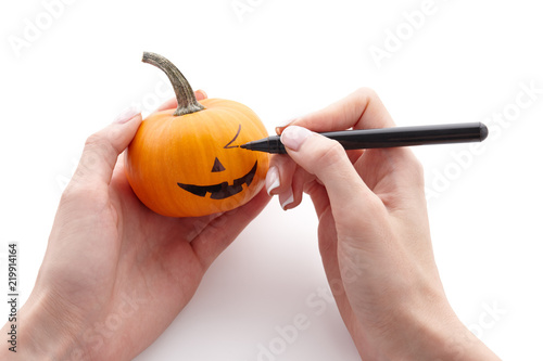 Woman's hands drawing scary face on little pumpkin with a black marker pen isolated on white background for Halloween. Holiday decoration concept. photo