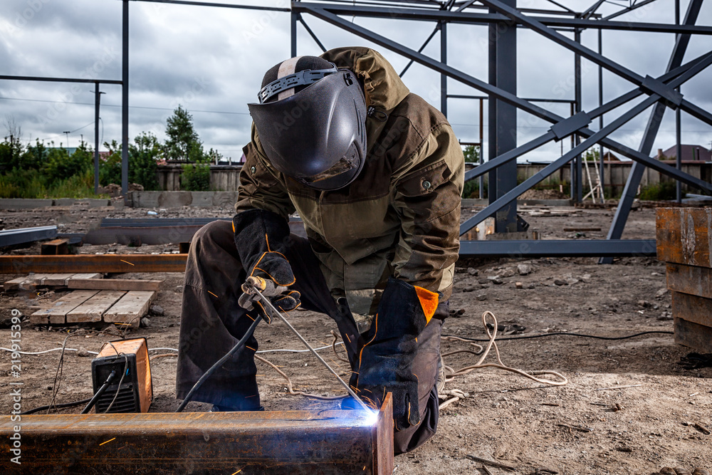 Man welder in welding mask, building uniform and blue protective gloves brews metal welding machine on street construction, in the background construction site Man welder in welding mask, building uniform and blue protective gloves brews metal welding machine on street construction, in the background construction site