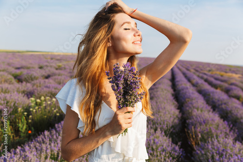 Photo of pretty european woman in dress holding bouquet with flowers, while walking outdoor through lavender field in summer
