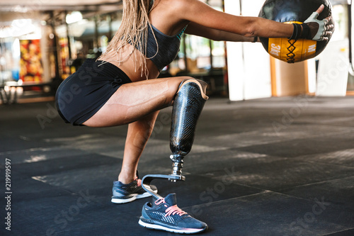 Portrait of beautiful invalid sportswoman with prosthesis in tracksuit, squatting with fitness ball in crossfit gym