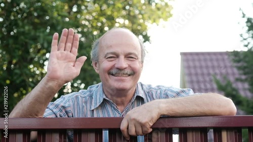 Friendly caucasian old man waving hi or farewell, isolated outdoors background with green trees and fence. Homeowmer saying goodbye to his guest