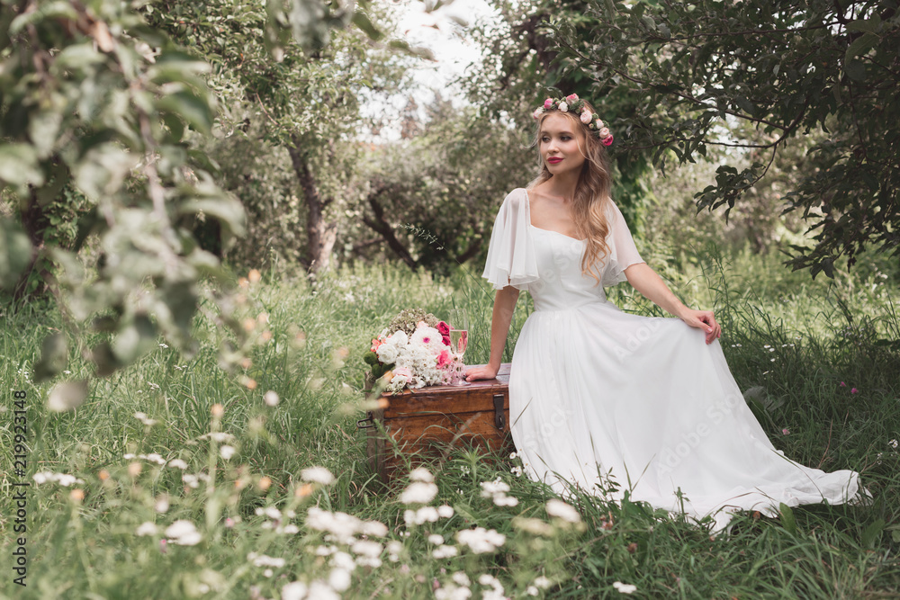 beautiful smiling young bride sitting on wooden chest and looking away in park beautiful smiling young bride sitting on wooden chest and looking away in park