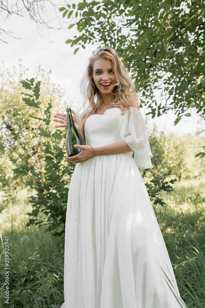 low angle view of young bride holding bottle of wine and smiling at camera low angle view of young bride holding bottle of wine and smiling at camera