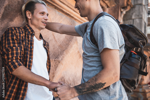 Optimistic man keeping hand of glad friend while situating near construction. They communicating while standing on street