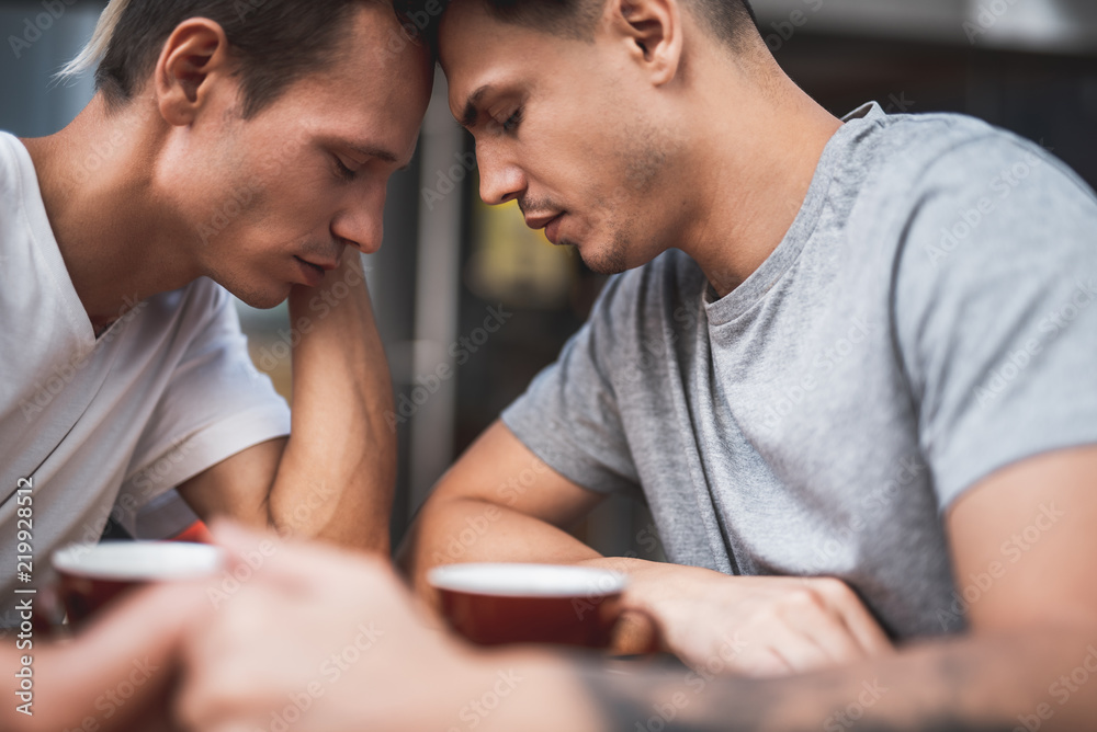 Side view calm male touching forehead of another serene guy while tasting cups of appetizing liquid at desk Side view calm male touching forehead of another serene guy while tasting cups of appetizing liquid at desk