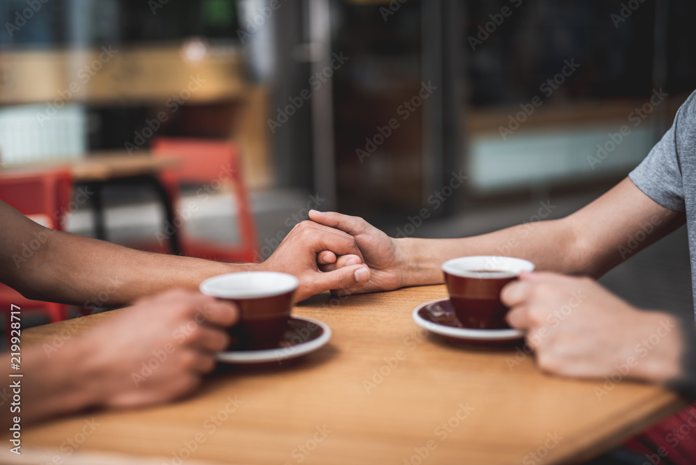 Close up two men keeping arms together during conversation while tasting mugs of beverage and locating at desk. Unity, love, brotherhood concept