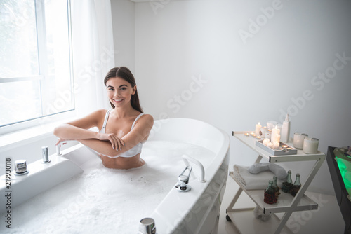 Portrait of satisfied lady taking bath while resting there. She looking at camera opposite window and candles