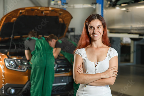 A beautiful girl poses for a photo while, two young mechanics in green overalls try to fix the engine of an orange car © Oleksandr