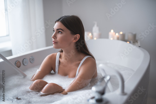 Portrait of thoughtful woman having bath with foam in apartment. Candles locating opposite her. Dreaming girl during rest concept