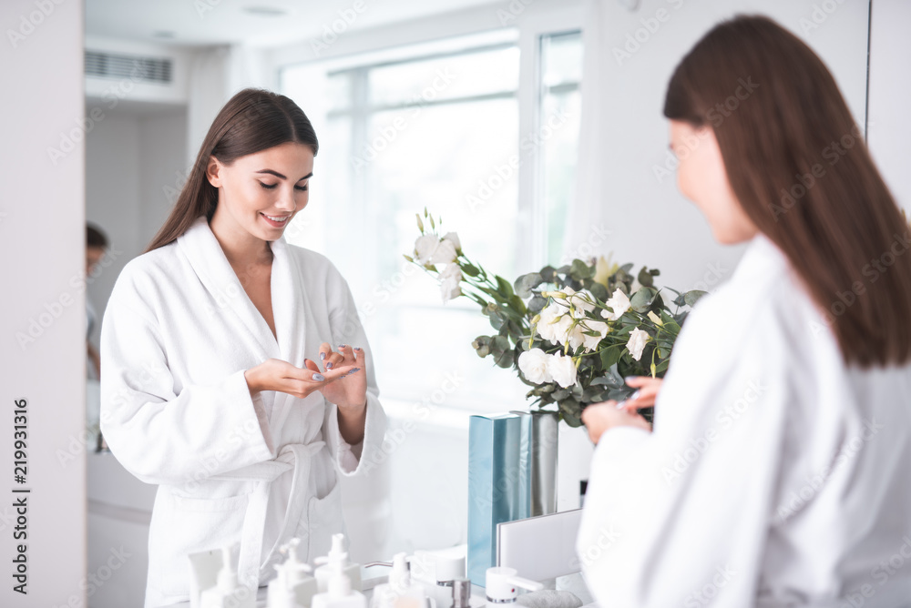 Portrait of happy woman keeping special liquid for taking care of skin on hands, while standing opposite mirror in bathrobe near flowers in white bright apartment Portrait of happy woman keeping special liquid for taking care of skin on hands, while standing opposite mirror in bathrobe near flowers in white bright apartment