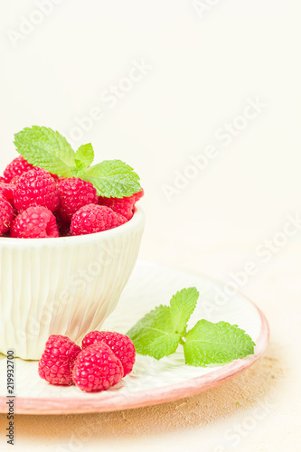 Ripe raspberries with green mint leaves in cup and saucer on pastel yellow background.