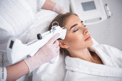 Automatic tool. Cosmetologist in sterile gloves using electronic syringe gun while lovely girl laying at daybed with closed eyes