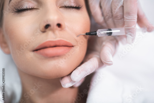 Enjoying skincare. Close up portrait of attractive girl with closed eyes and cosmetologist hands with syringe. Doctor carefully holding woman face