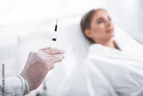 Ready for procedure. Close up of cosmetologist arm holding needle. Woman in soft bathrobe lying on deckchair