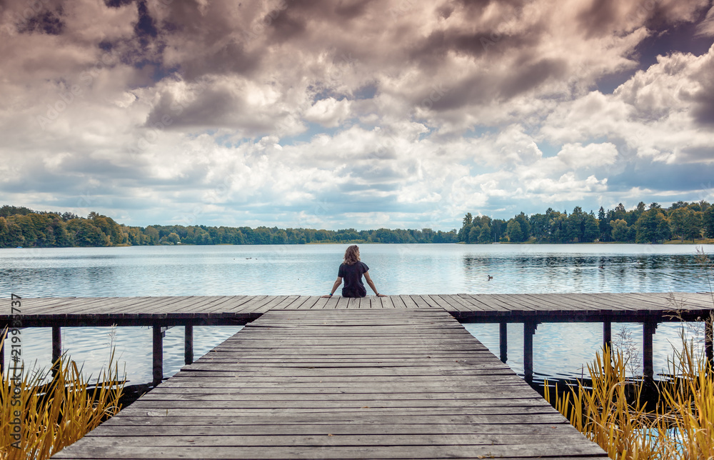 Girl woman sits with her back on a wooden pier and looks at the landscape, lake and clouds, Trakai, Lithuania