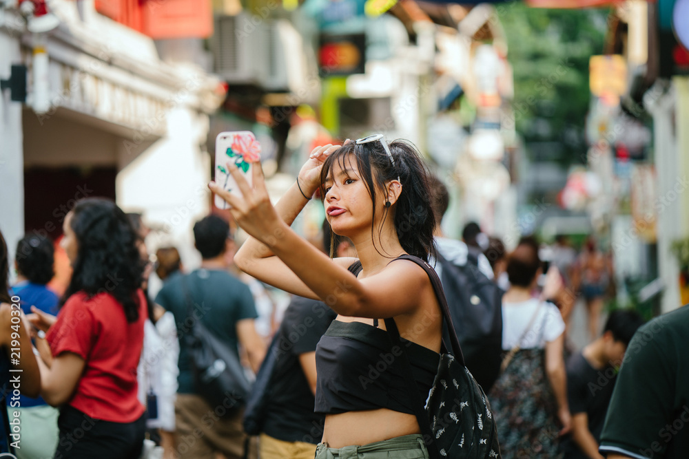 Portrait of a young Chinese millennial young girl taking a selfie of herself along a colorful and busy street in Asia at daytime She is wearing trendy street clothes. Portrait of a young Chinese millennial young girl taking a selfie of herself along a colorful and busy street in Asia at daytime She is wearing trendy street clothes.