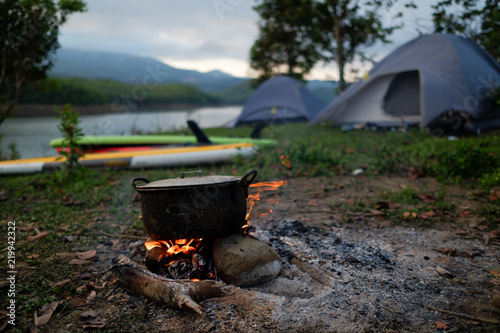 Fototapeta Naklejka Na Ścianę i Meble -  Camp and cooking in field conditions, boiling pot at the campfire on picnic in morning.  Cooking dinner on firewood stove using firewood when going to the wilderness or outdoor activity, camping tent