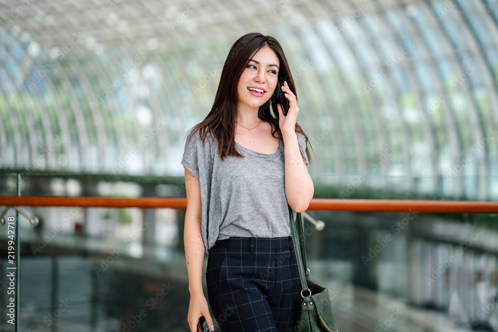 A young and attractive Japanese Asian woman is having a conversation on her smartphone. She is smiling as she speaks on her phone and is standing in a futuristic and modern building.