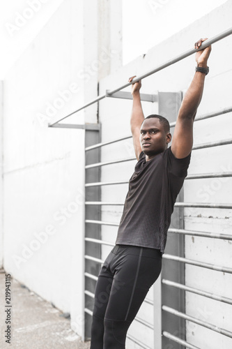 Concentrated african american sportsman warming up and stretching legs on pier, pulls up on a horizontal bar