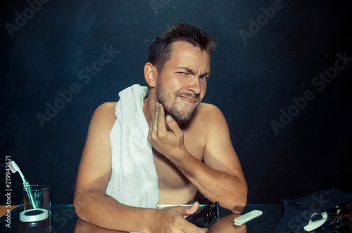 The young man in bedroom sitting in front of the mirror scratching his beard at home. Human emotions concept