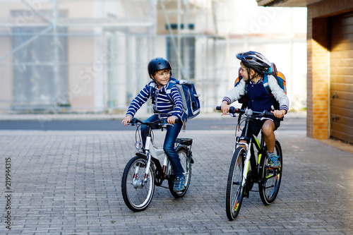 Two school kid boys in safety helmet riding with bike in the city with backpacks. Happy children in colorful clothes biking on bicycles on way to school. Safe way for kids outdoors to school