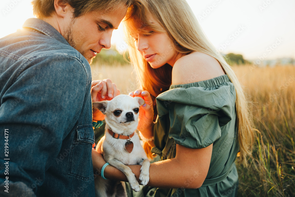 A young couple are sitting on a yellow autumn grass and hugging their white chihuahua breed dog. A young couple are sitting on a yellow autumn grass and hugging their white chihuahua breed dog.