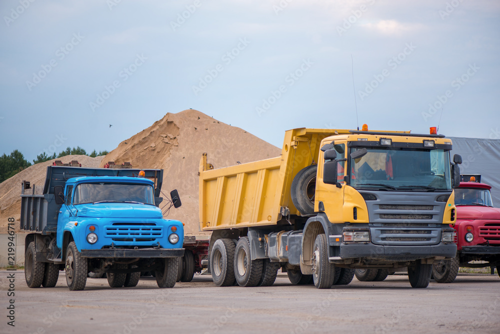 Multiple cars, trucks, loaders, concrete mixers and construction machinery in large parking lot in industrial territory, next to concrete and asphalt factory. Raw material heaps, gravel in background