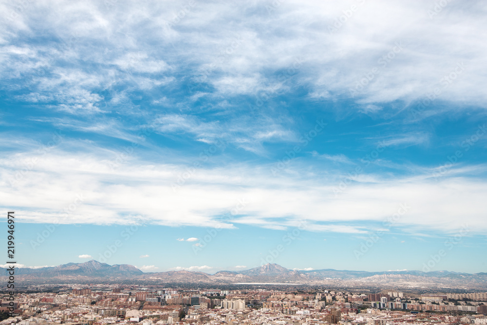 Panoramic view of city from Santa Barbara Castle in Alicante, Spain. Block apartment buildings, parks, roads, houses, palm trees. Beautiful mountain landscape in background, blue sky 