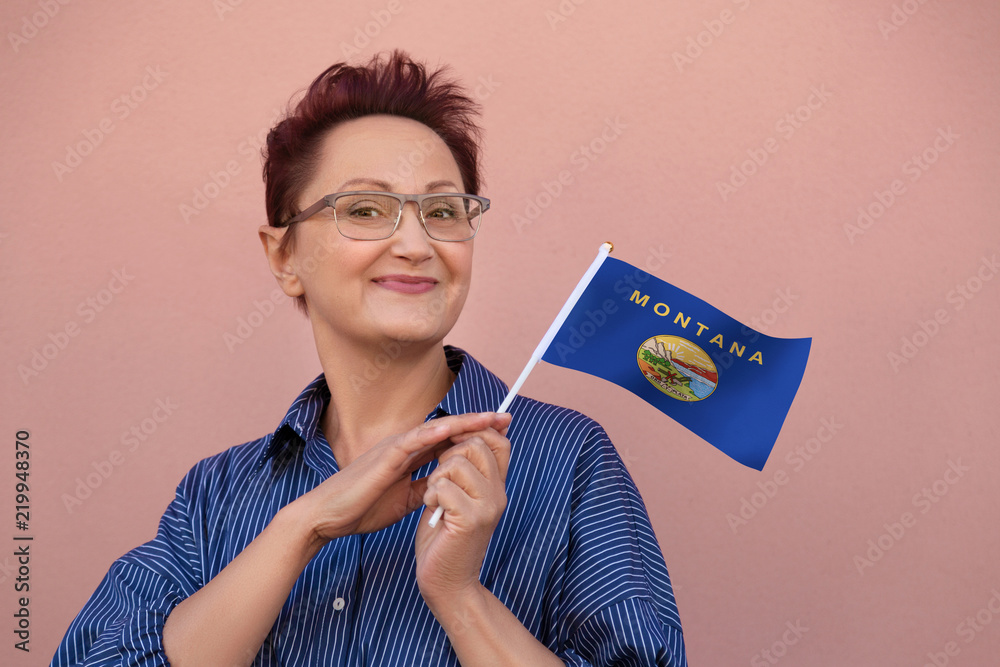 Montana flag. Woman holding Montana state flag. Nice portrait of middle aged lady 40 50 years old with a state flag over pink wall on the street outdoors. Montana flag. Woman holding Montana state flag. Nice portrait of middle aged lady 40 50 years old with a state flag over pink wall on the street outdoors.