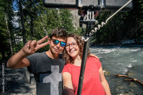 Couple of friends taking self portrait with selfie stick with a mountain river in background. Two young brothers make selfie on a phone outside in the park. Teens taking a self picture with smartphone