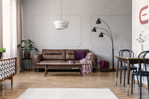 Real photo of light grey sitting room interior with brown couch with blanket and violet pillow, coffee table with two tea cups, metal lamp and molding on wall photo