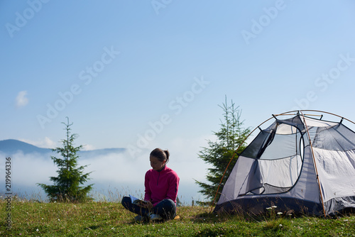 Back view of young woman hiker sitting on green grass of blooming valley at tourist tent under beautiful blue sky reading a book on bright summer morning on foggy mountains background.
