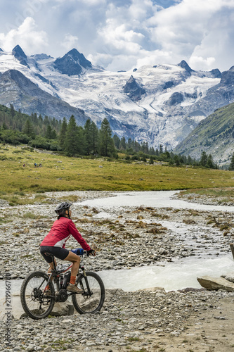 active senior woman, riding her e-mountain bike in the Roseg valley below the glaciers and summits of the Sella Group and Piz Roseg