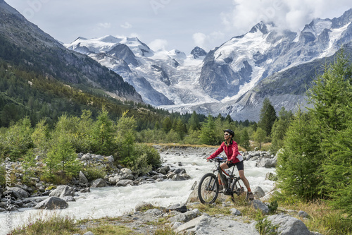 Senior woman, riding her e-mountain bike on the trail to the Morteratsch glacier near Pontresina , Engadin, Switzerland,Alps. In the background the famous mountains of Piz palu, Bellavista, Bernina
