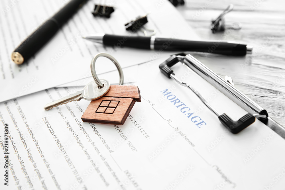 Key with trinket in shape of house and mortgage contract on table, closeup Key with trinket in shape of house and mortgage contract on table, closeup