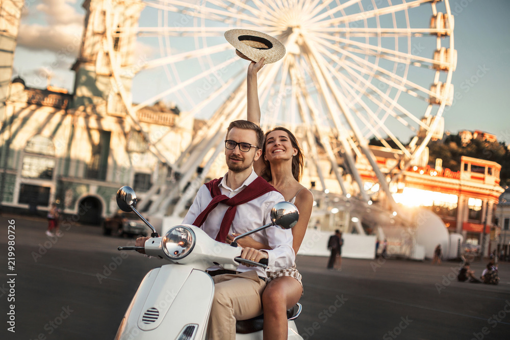 Husband and wife ride around the city on a white motor scooter in the background of a ferris wheel