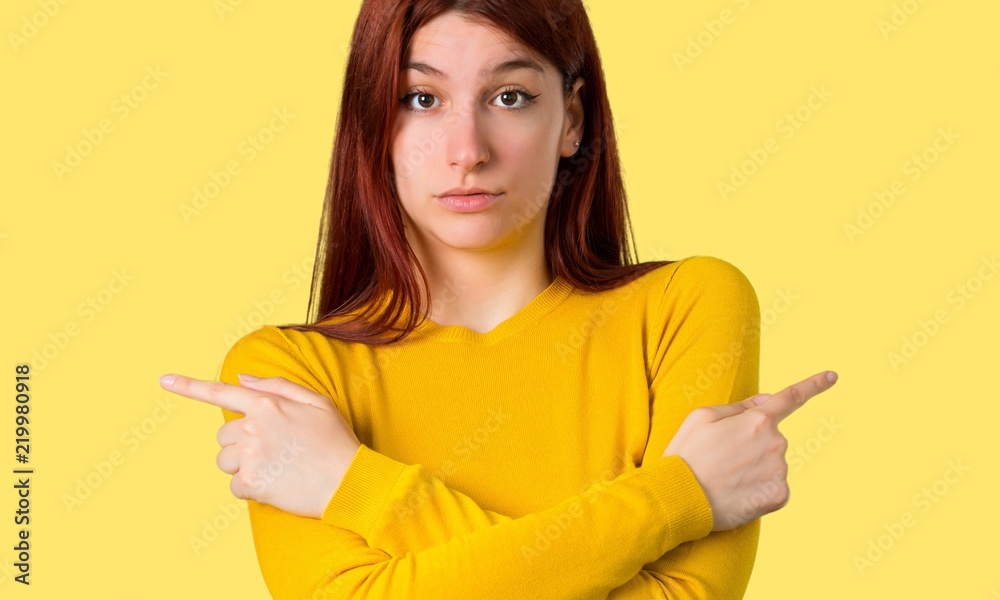 Young redhead girl with yellow sweater pointing to the laterals having doubts. Undecided person on isolated yellow background Young redhead girl with yellow sweater pointing to the laterals having doubts. Undecided person on isolated yellow background