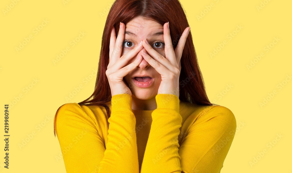 Young redhead girl with yellow sweater surprised and covering face with hands while looking through fingers on isolated yellow background Young redhead girl with yellow sweater surprised and covering face with hands while looking through fingers on isolated yellow background